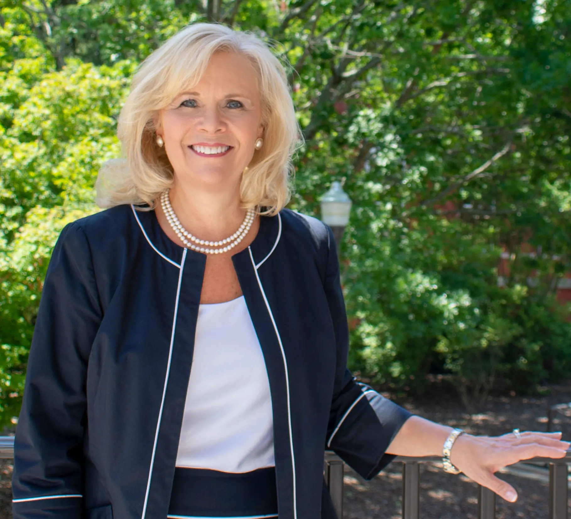 Susan Hubbard smiles outdoors in front of green trees, wearing a dark jacket with white trim and a pearl necklace.