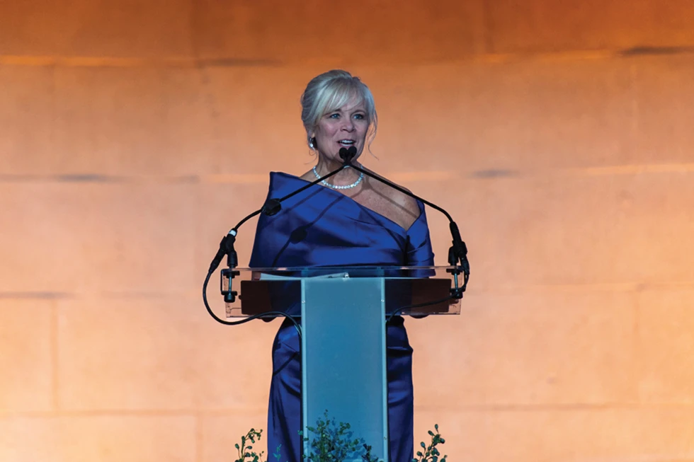 Woman in a blue formal gown speaks at a podium during an indoor event.