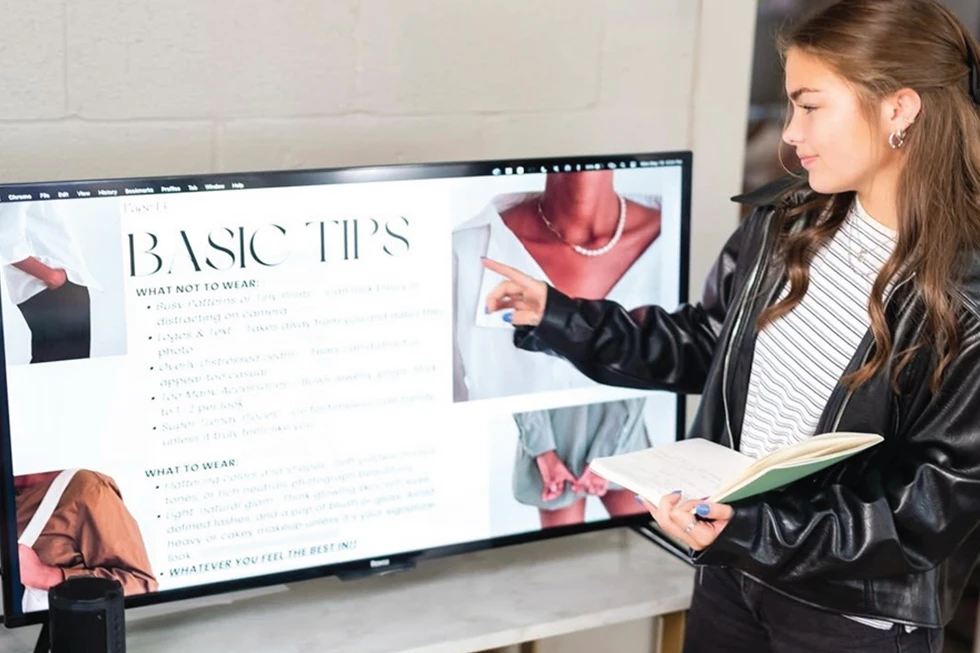 Student points to a screen displaying fashion styling tips while holding an open notebook.
