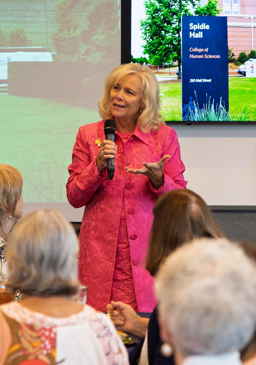 Dean Susan Hubbard, wearing a pink jacket, speaks into a handheld microphone during the luncheon. A screen displaying Spidle Hall is visible in the background.