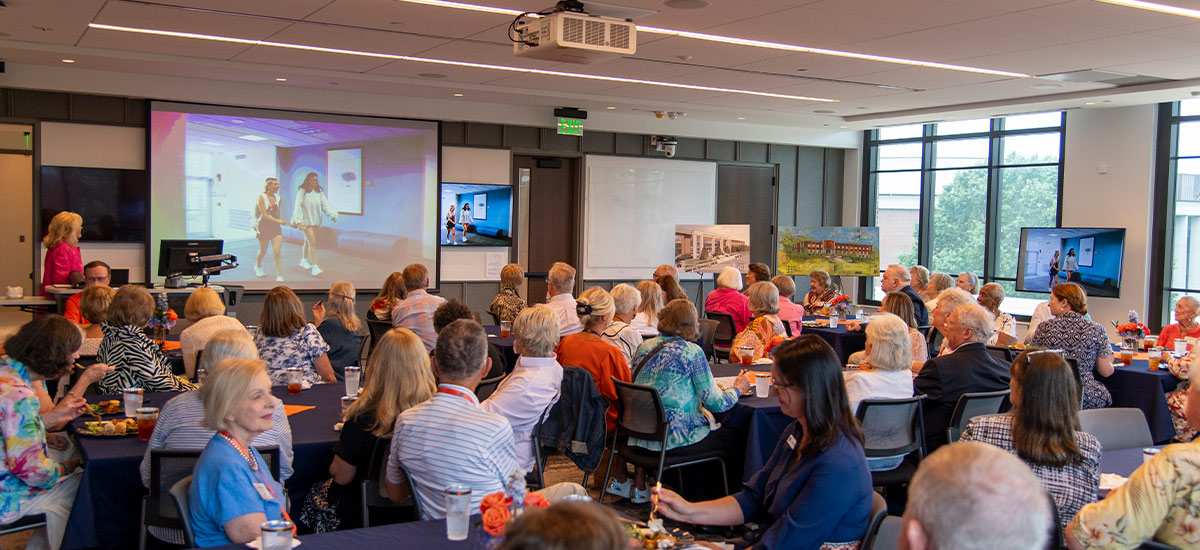 Alumni sit at tables in a large room during the Golden Eagles luncheon, facing a projector screen displaying a presentation.
