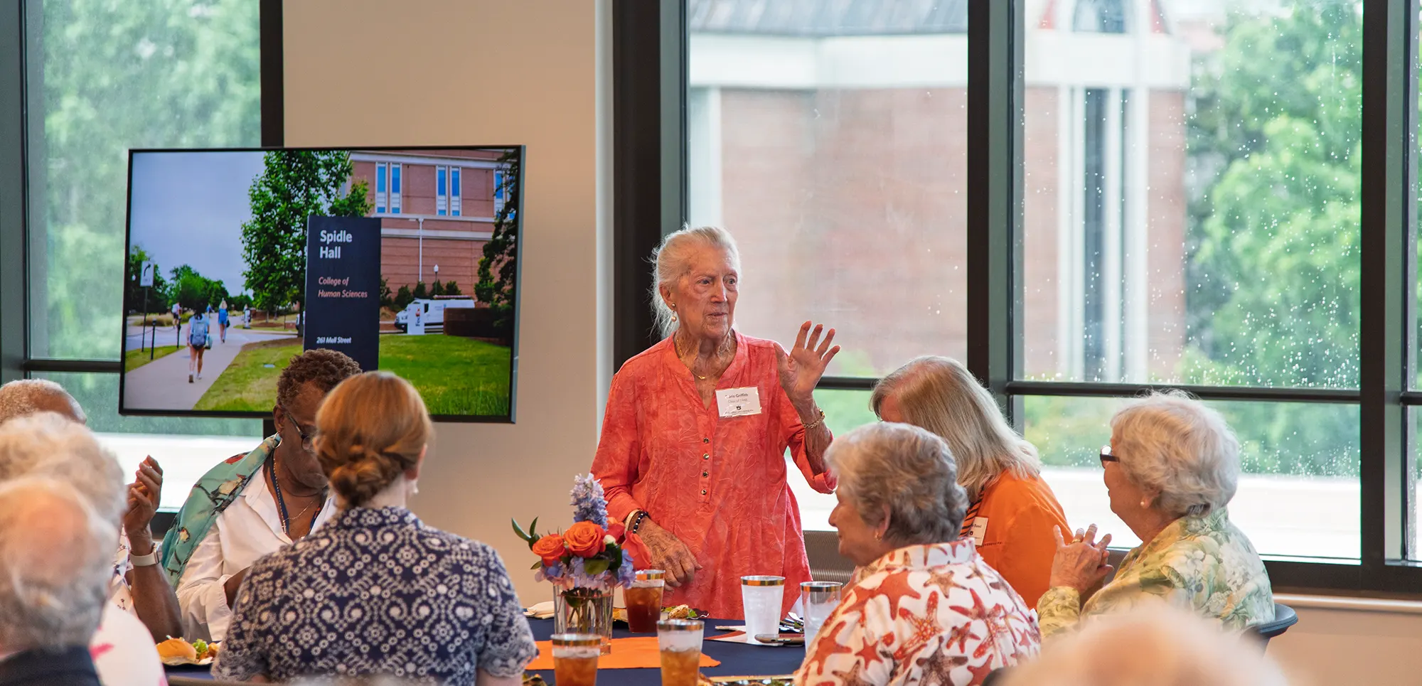 Doris Griffith, a 1948 alumna, stands to speak to fellow alumni at a luncheon table. A monitor behind her displays a photo of Spidle Hall.