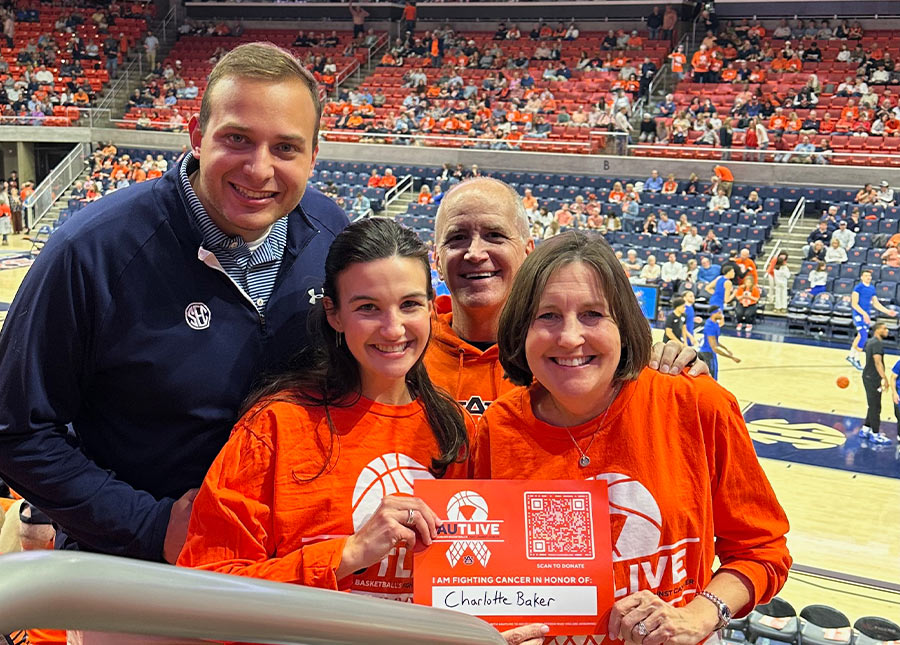 The Baker family smiling in the stands at an Auburn basketball game, holding a red AUTLIVE sign that reads, "I am fighting cancer in honor of: Charlotte Baker."