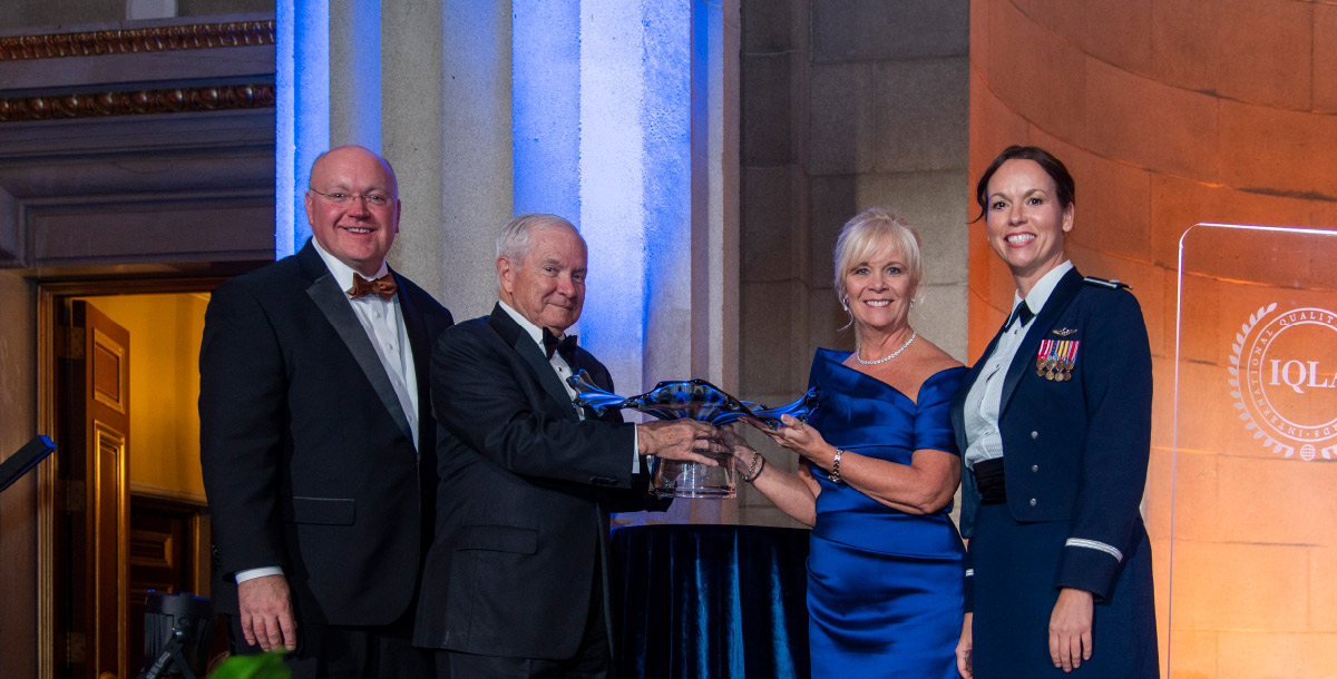 Four people on stage, including a woman in military uniform, presenting a glass award during a formal ceremony.