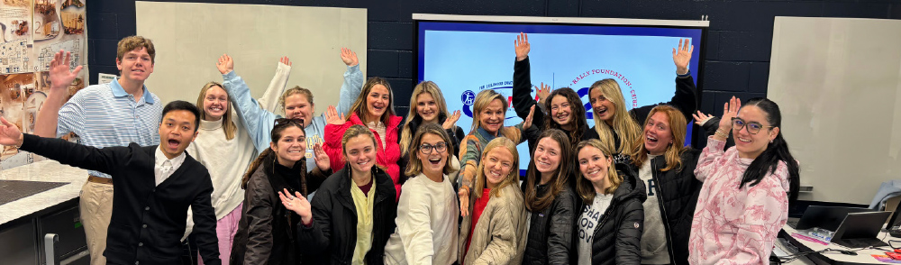 Large group of happy students and a speaker waving for a photo in front of a Rally Foundation digital display in a classroom.