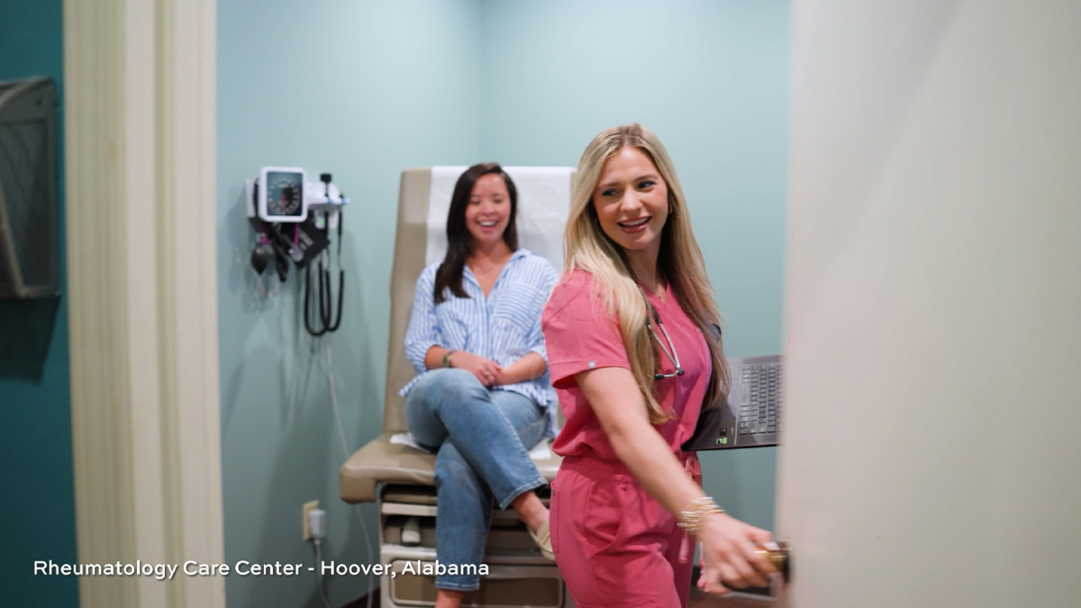 A female medical professional in pink scrubs enters an exam room to greet a seated patient at Rheumatology Care Center in Hoover, Alabama.