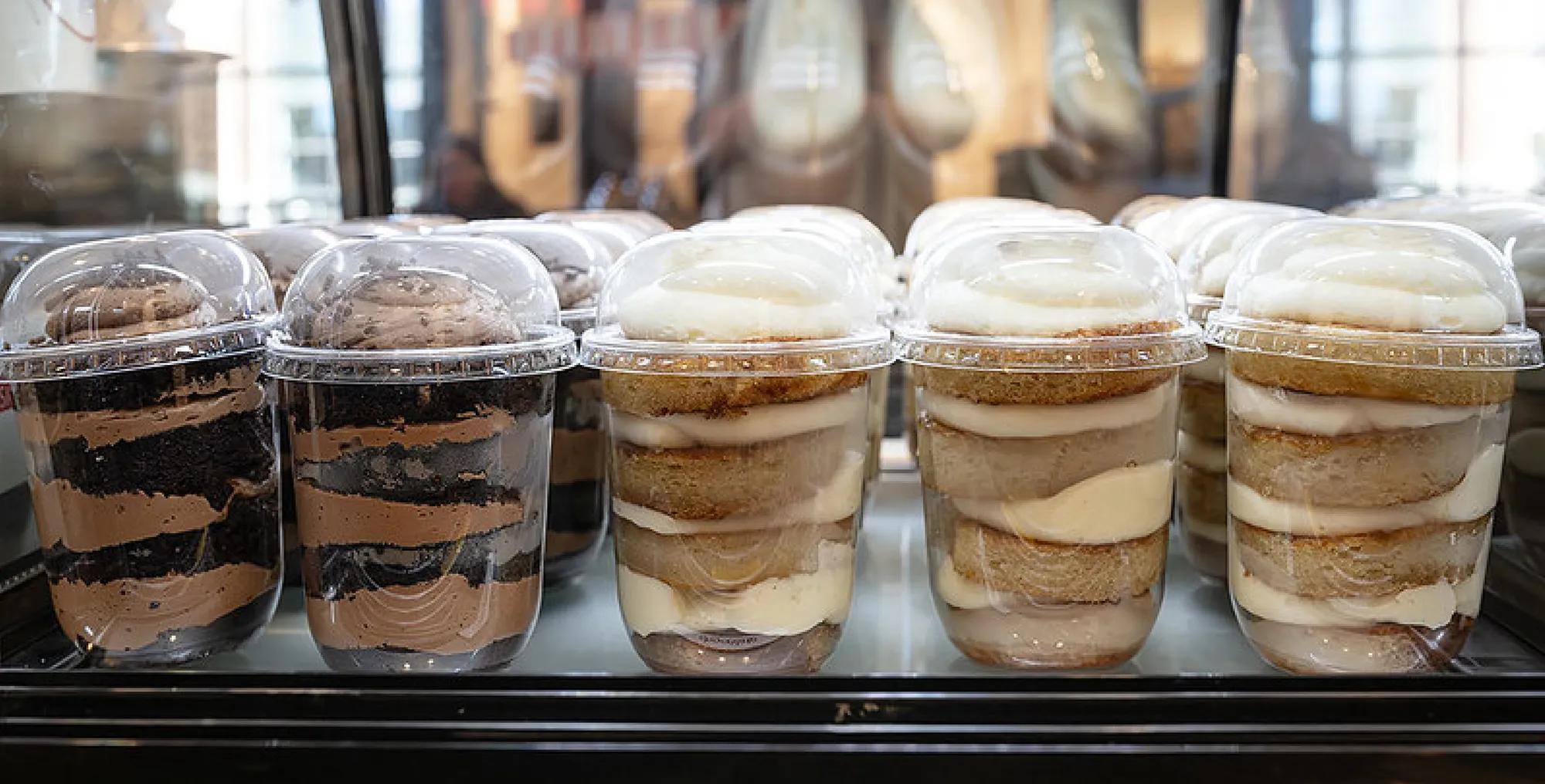 A row of clear plastic cups filled with layered cake and frosting parfaits. Varieties include chocolate with dark frosting and vanilla with white cream frosting, topped with domes.