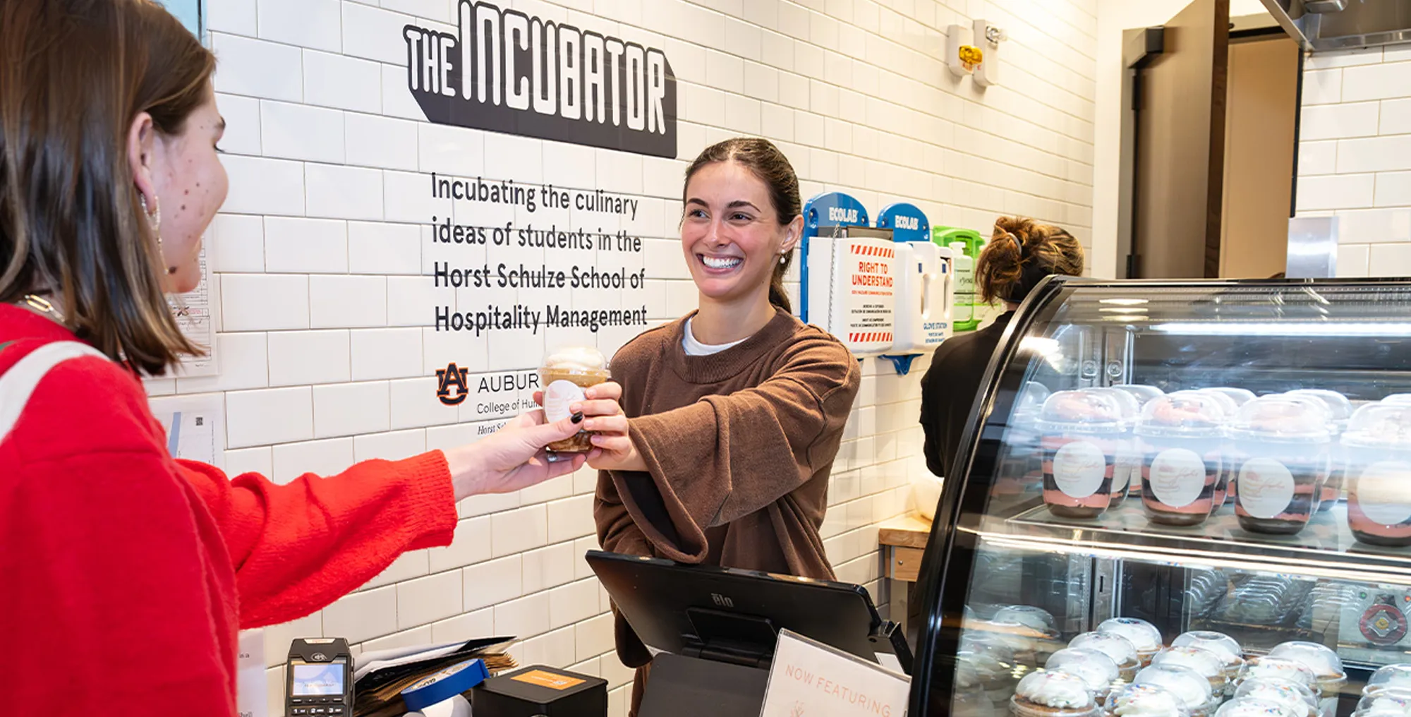 A smiling woman hands a layered cake cup to a customer over a counter at "The Incubator." A refrigerated display case filled with dessert cups is visible in the foreground.