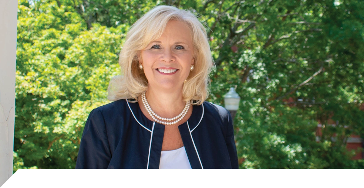 A smiling blonde woman in a dark blue jacket with white trim and pearl necklace, standing before a backdrop of green trees.
