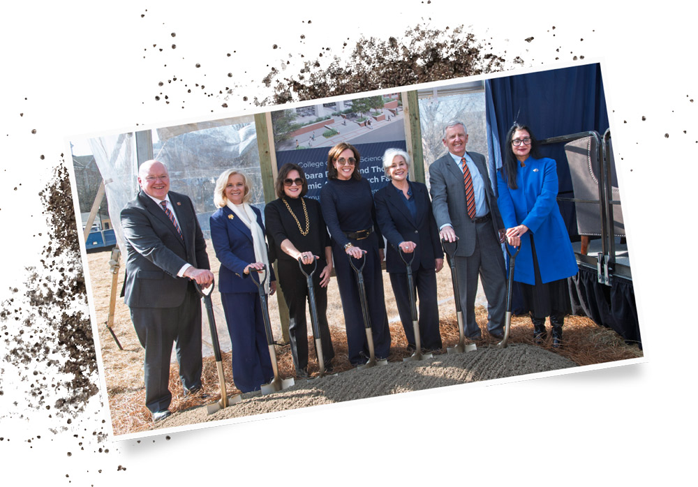 A group of seven professionally dressed individuals stand in a row behind a pile of dirt, each holding a ceremonial shovel during an outdoor university groundbreaking event.