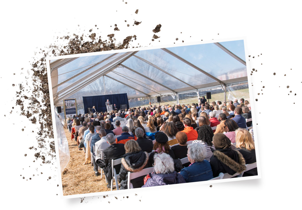 A wide view from the back of a large crowd seated in white chairs under a clear event tent, watching speakers on a stage during a formal outdoor university ceremony.