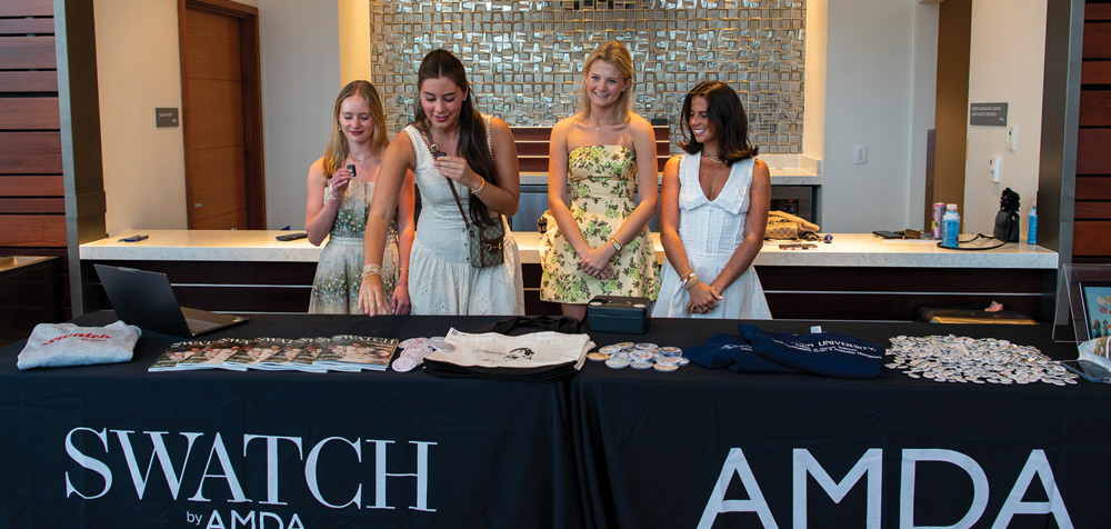 Four young women stand behind a black branded table labeled "SWATCH by AMDA," examining items for an event display.