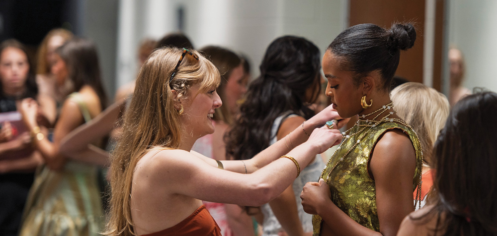 A woman adjusts a necklace on another person wearing a textured gold dress, preparing for a fashion show backstage.