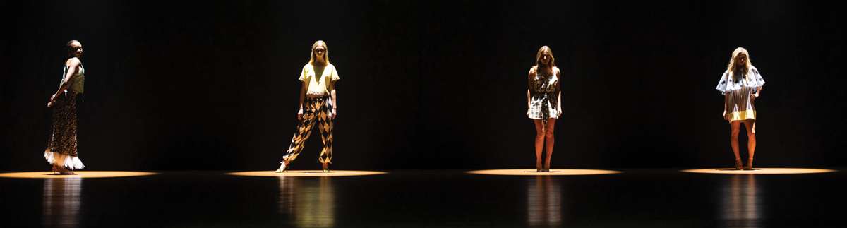 Four models stand separately in bright spotlights on a dark runway during a fashion show presentation.