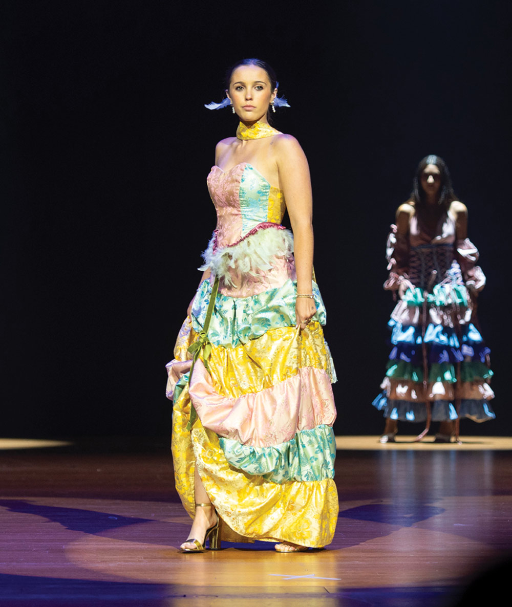 A model in a large, colorful, tiered patterned dress walks down a runway, with another model visible behind her.