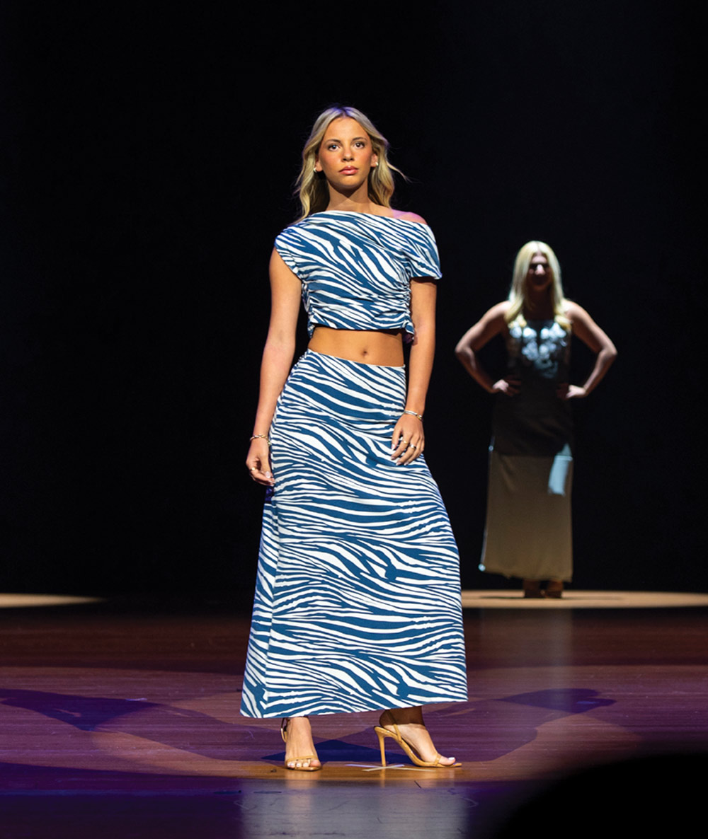 A model in a blue and white zebra-print two-piece outfit walks down a runway, with another model waiting behind.
