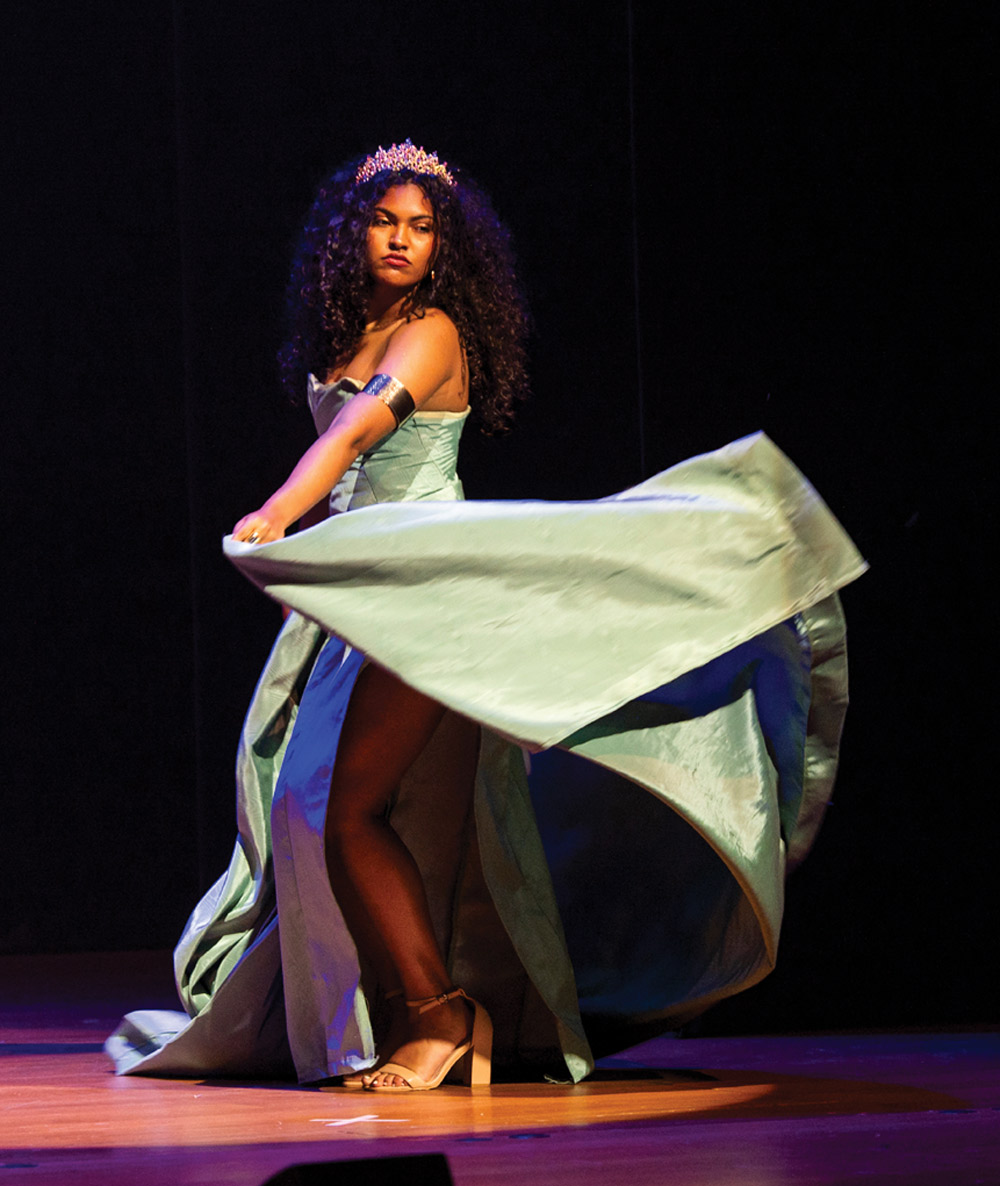 A model with curly hair, wearing a crown and a light blue flowing dress, poses dramatically on stage while turning.