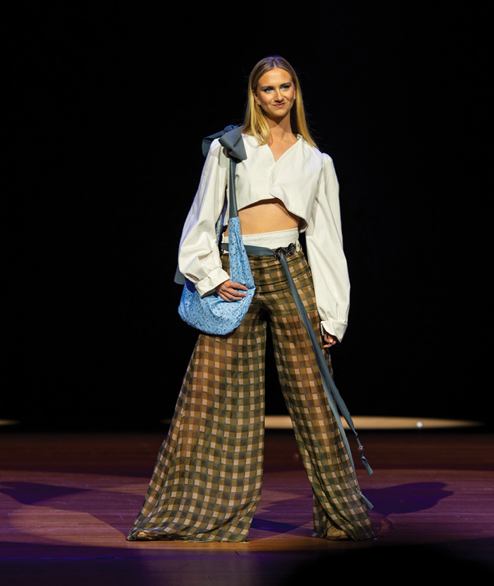 A model in a white crop top and brown plaid wide-leg pants carries a blue bag while walking down the runway.