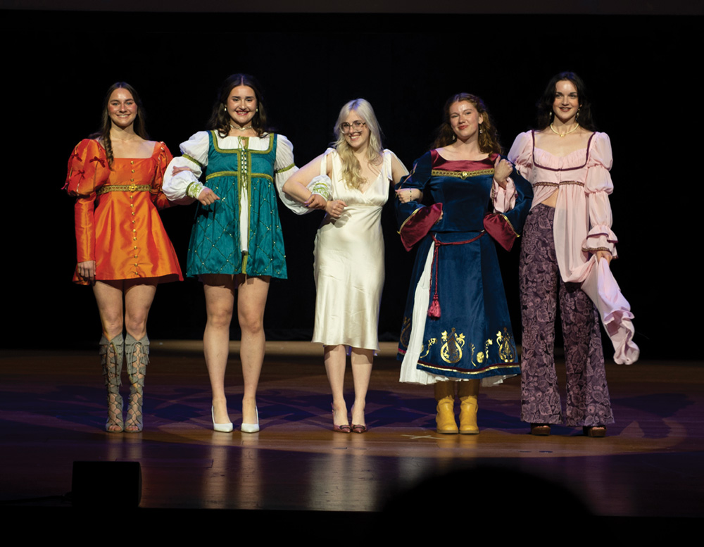 Five models stand in a line on stage, holding hands, wearing diverse and colorful outfits during a fashion show.