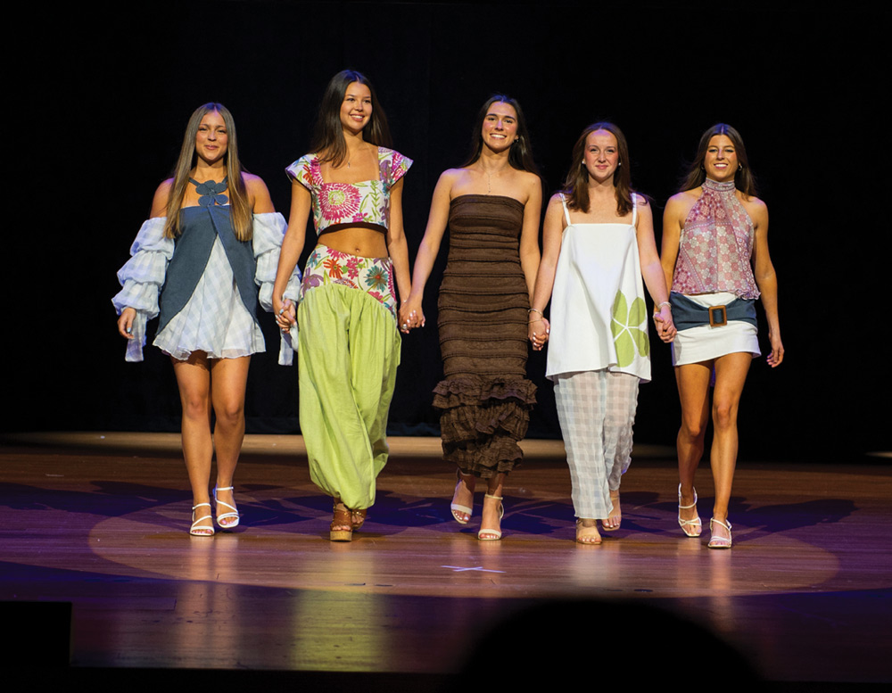 Five models walk hand-in-hand across the stage during a fashion show, wearing stylish, colorful casual outfits.