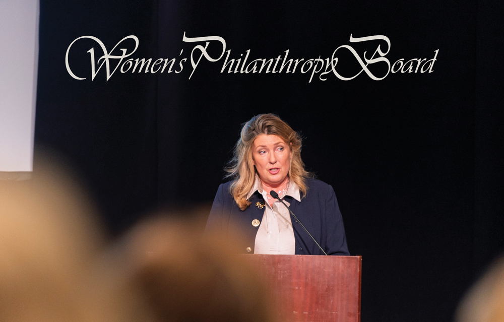 A woman in a dark blazer speaks from a podium at an event in front of a "Women's Philanthropy Board" sign.
