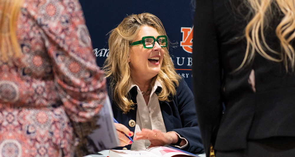 A smiling woman wearing bright green glasses signs a book while interacting with people at an event.