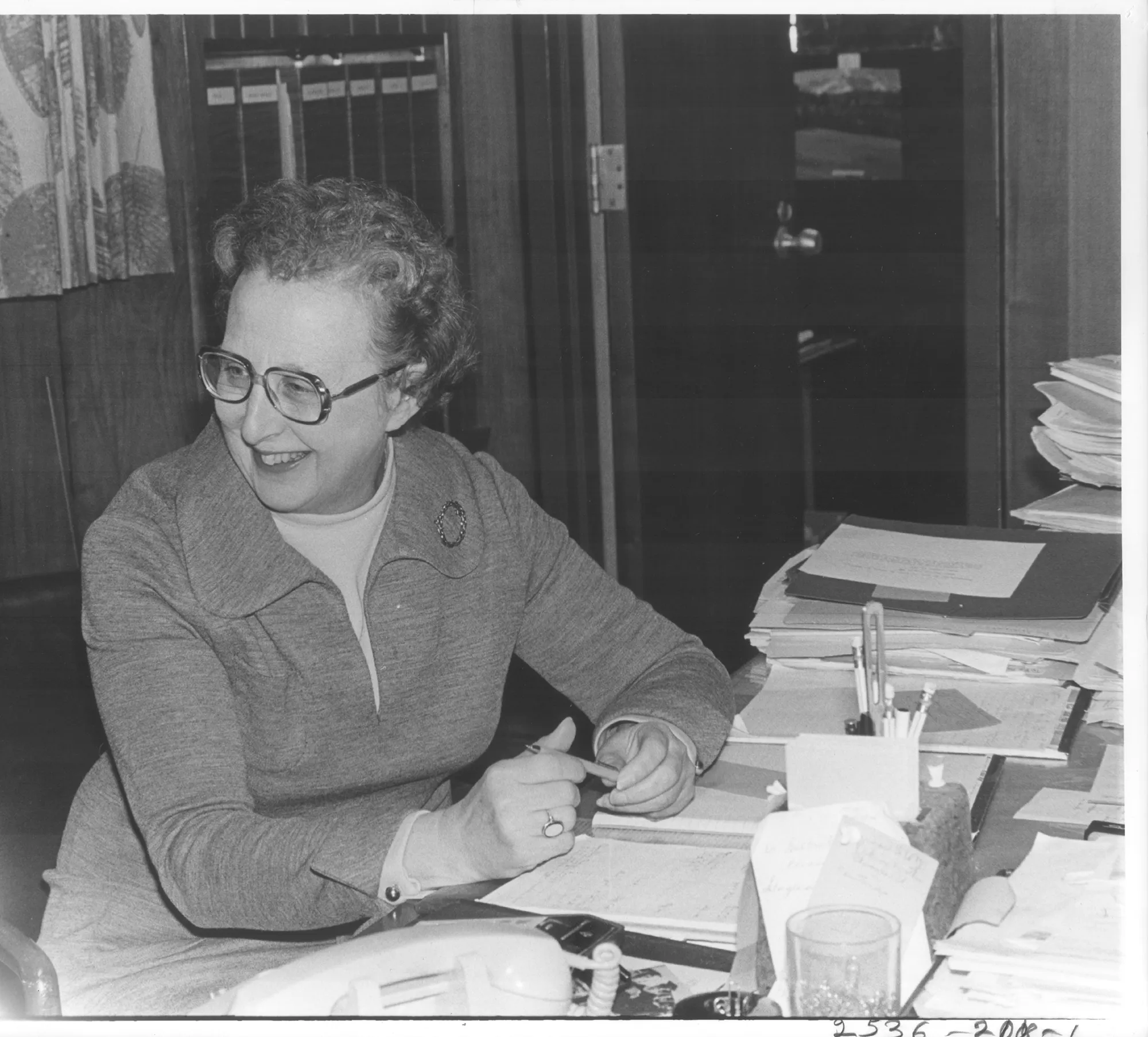 A vintage black-and-white photo of a smiling woman with glasses sitting at a desk cluttered with papers, folders, and a rotary phone, appearing to be in the middle of a workday.