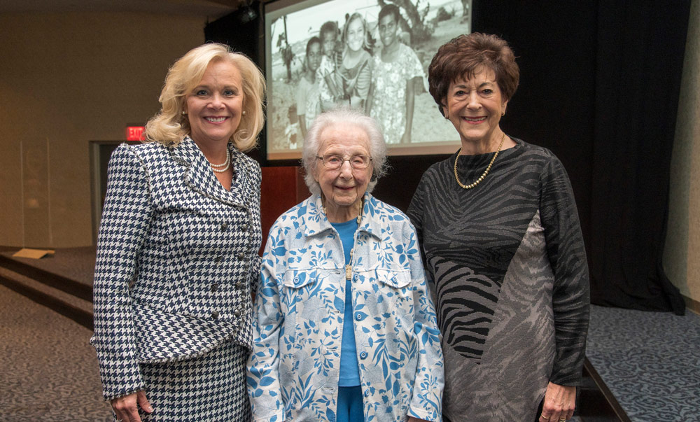 Three women smile for a photo at an event. They stand in front of a projection screen showing a historical image of children, with the woman in the center wearing a blue floral jacket.