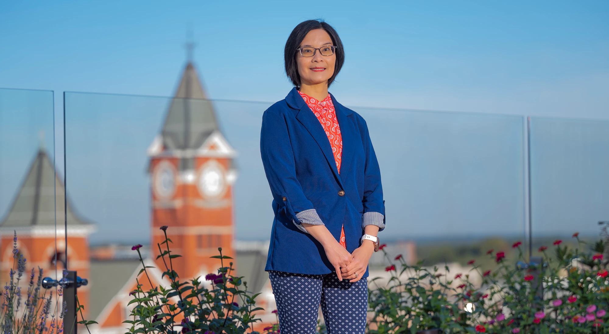 A woman in a blue blazer and red patterned shirt stands on a rooftop garden. Behind her, the iconic orange brick clock tower of Auburn University is visible against a clear blue sky.