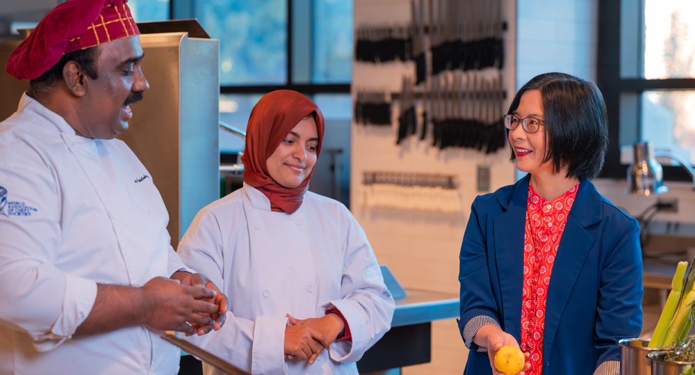 In a commercial kitchen, a woman in a blue blazer talks with two chefs in white uniforms. She holds a lemon while engaging in a friendly conversation near a stainless steel counter.