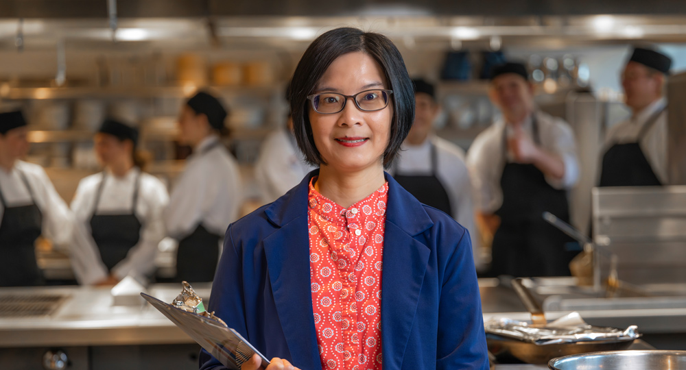 A professional portrait of a woman wearing glasses and a blue blazer, holding a clipboard in a commercial kitchen. In the blurred background, several chefs work at their stations.