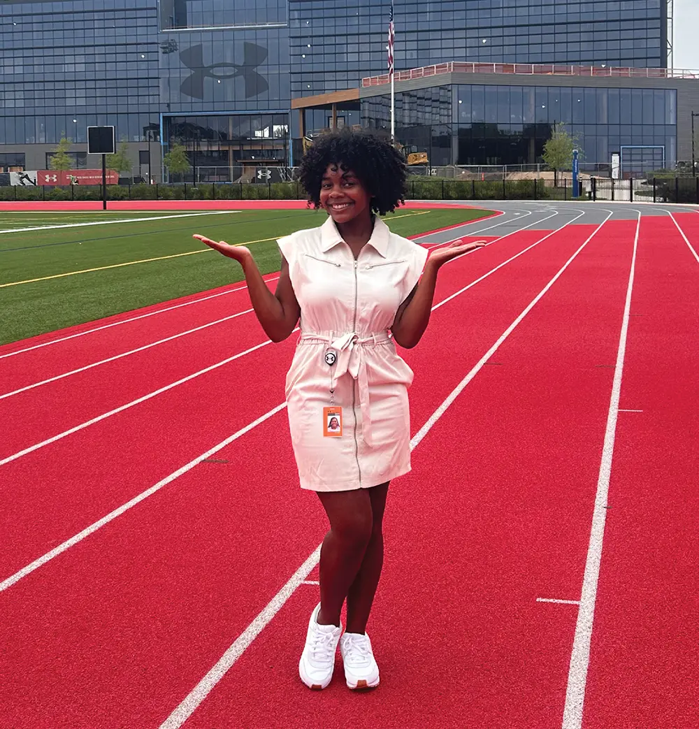 A woman with curly hair smiling on a red running track, with a large glass building featuring an Under Armour logo in the back