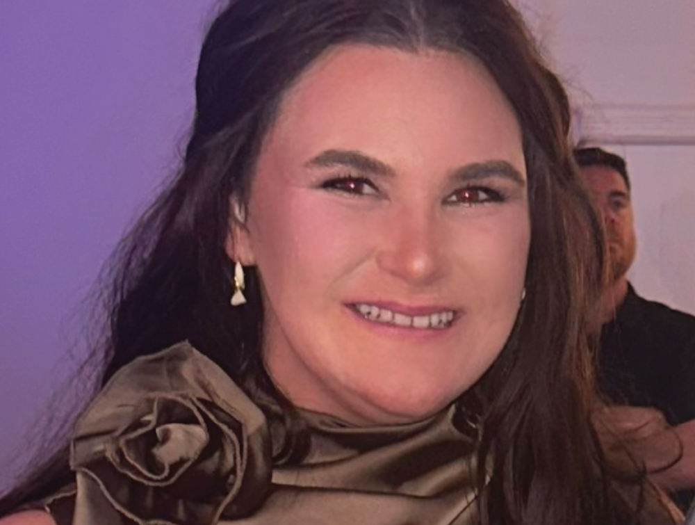 A smiling woman with long dark hair wearing a bronze-colored ruffled top in a close-up portrait shot.