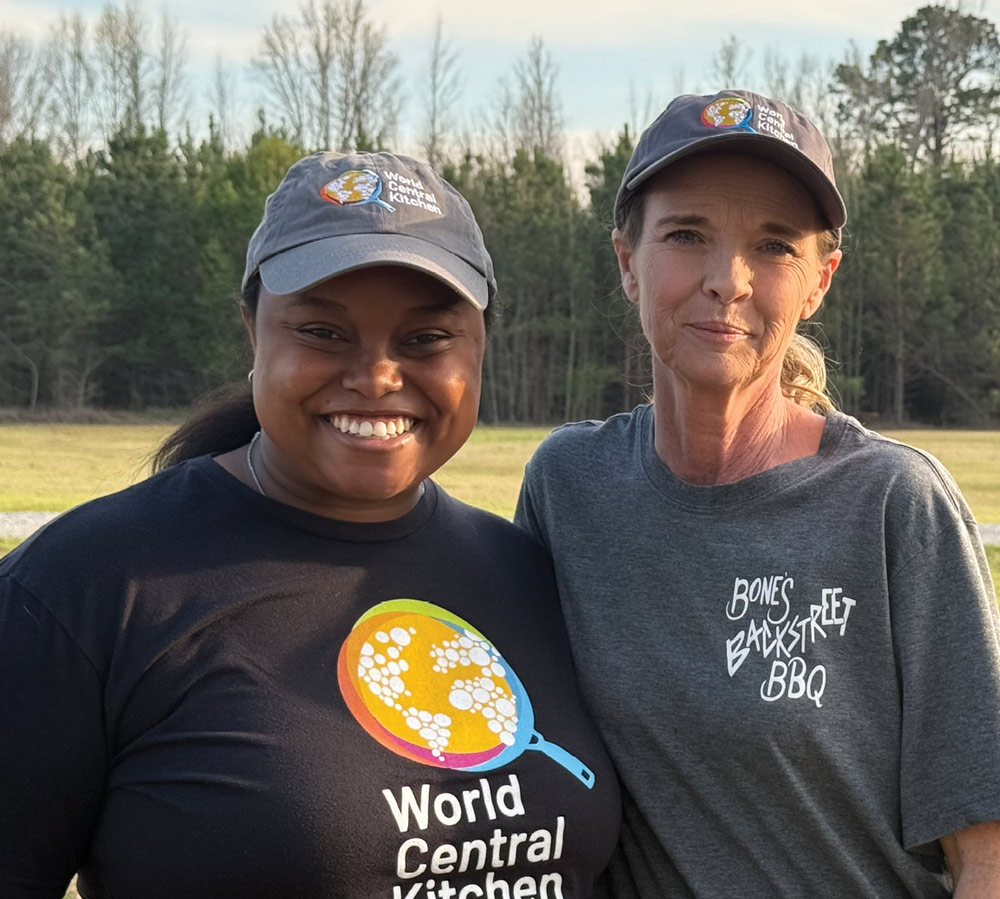 Two women smiling side-by-side, both wearing gray World Central Kitchen caps, outdoors in front of a tree line.