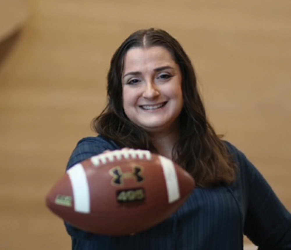 A woman smiling while holding an Under Armour branded football out toward the camera in a close-up shot.