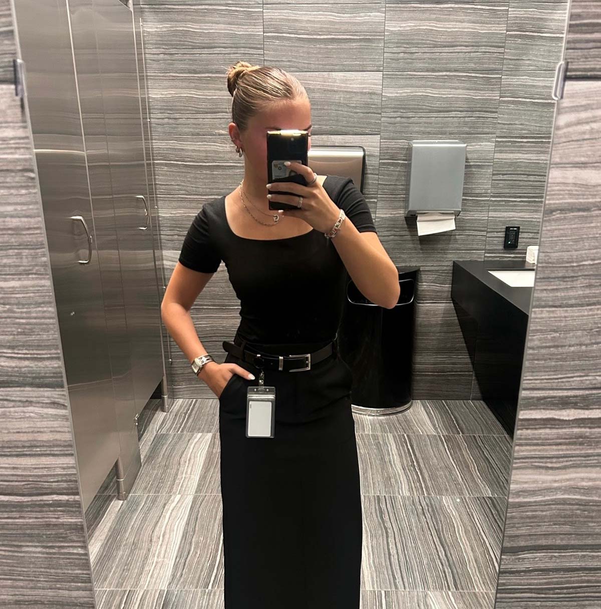 A mirror selfie of a woman with hair in a bun, wearing an all-black outfit including a square-neck top, pants, and a belt with an attached ID badge