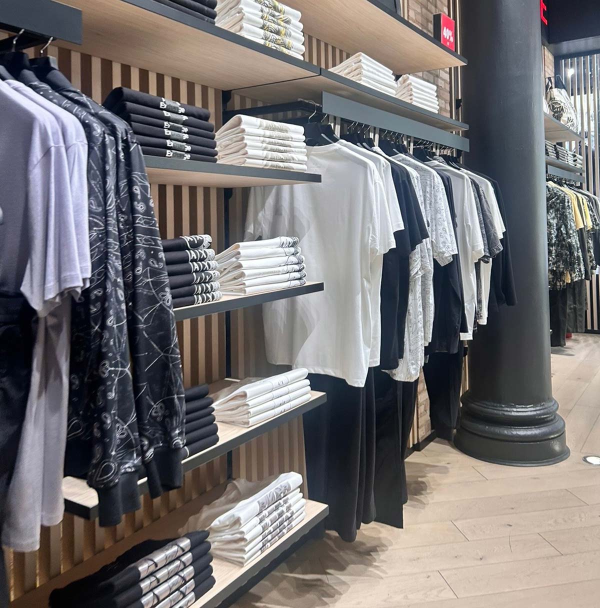 Interior of a retail clothing store featuring neatly folded black and white t-shirts on wooden shelves and apparel hanging on racks against a modern slatted wall