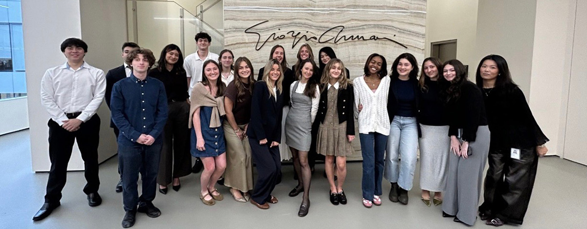 A large group of smiling young people pose for a photo in front of a stone wall with the signature script for 'Giorgio Armani'