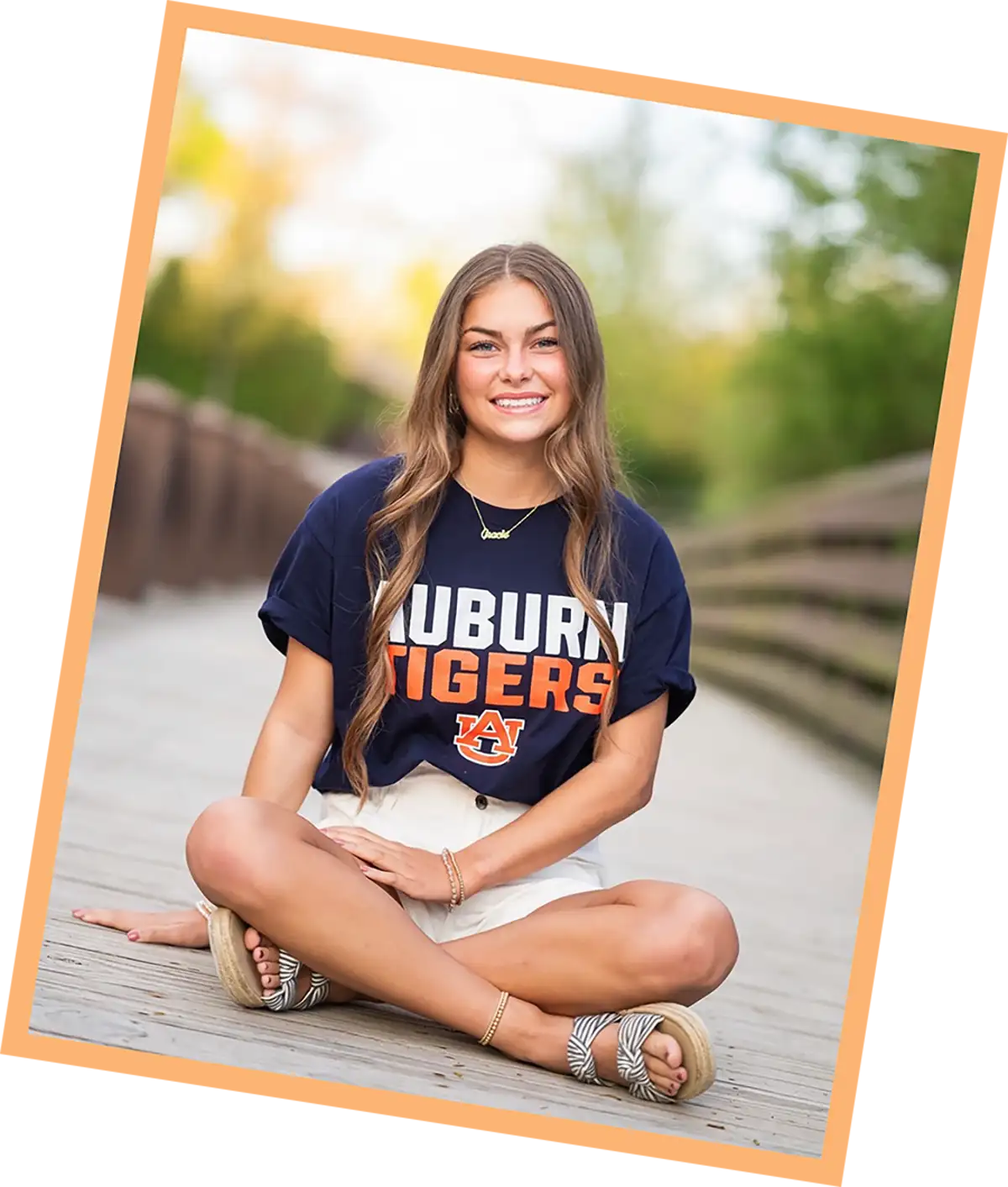 A tilted, peach-bordered photo of a smiling young woman sitting cross-legged on a wooden bridge, wearing a navy Auburn Tigers t-shirt and white shorts
