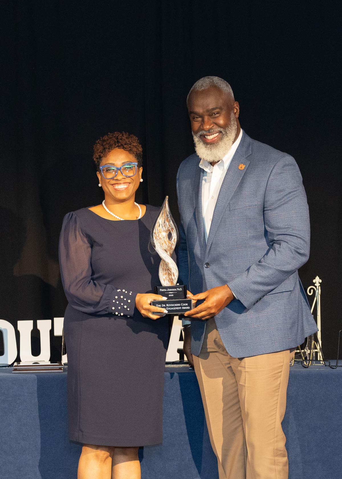 A smiling woman with blue glasses and a dark blue dress receives a glass, flame-shaped award from a smiling man with a white beard wearing a blue patterned blazer and khaki pants