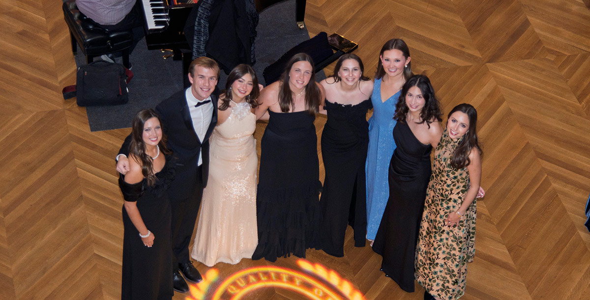 High-angle view of eight young adults in formal evening gowns and a tuxedo posing on a geometric patterned wooden floor