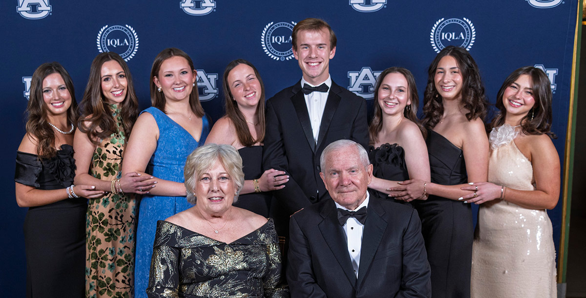 A formal group photo of an older couple seated in front of eight young adults in evening wear, posing against a blue Auburn University and IQLA backdrop.