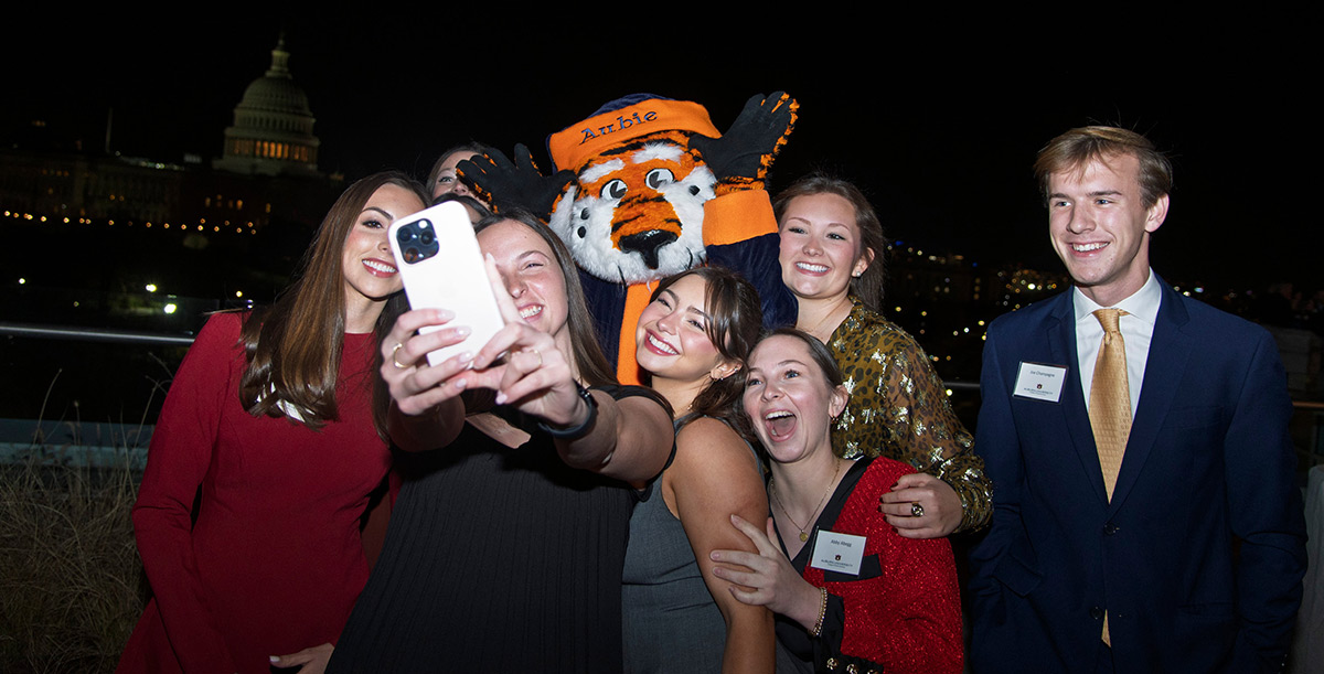 A group of smiling young adults and the Auburn mascot, Aubie the Tiger, take a selfie at night with the illuminated US Capitol building in the background.