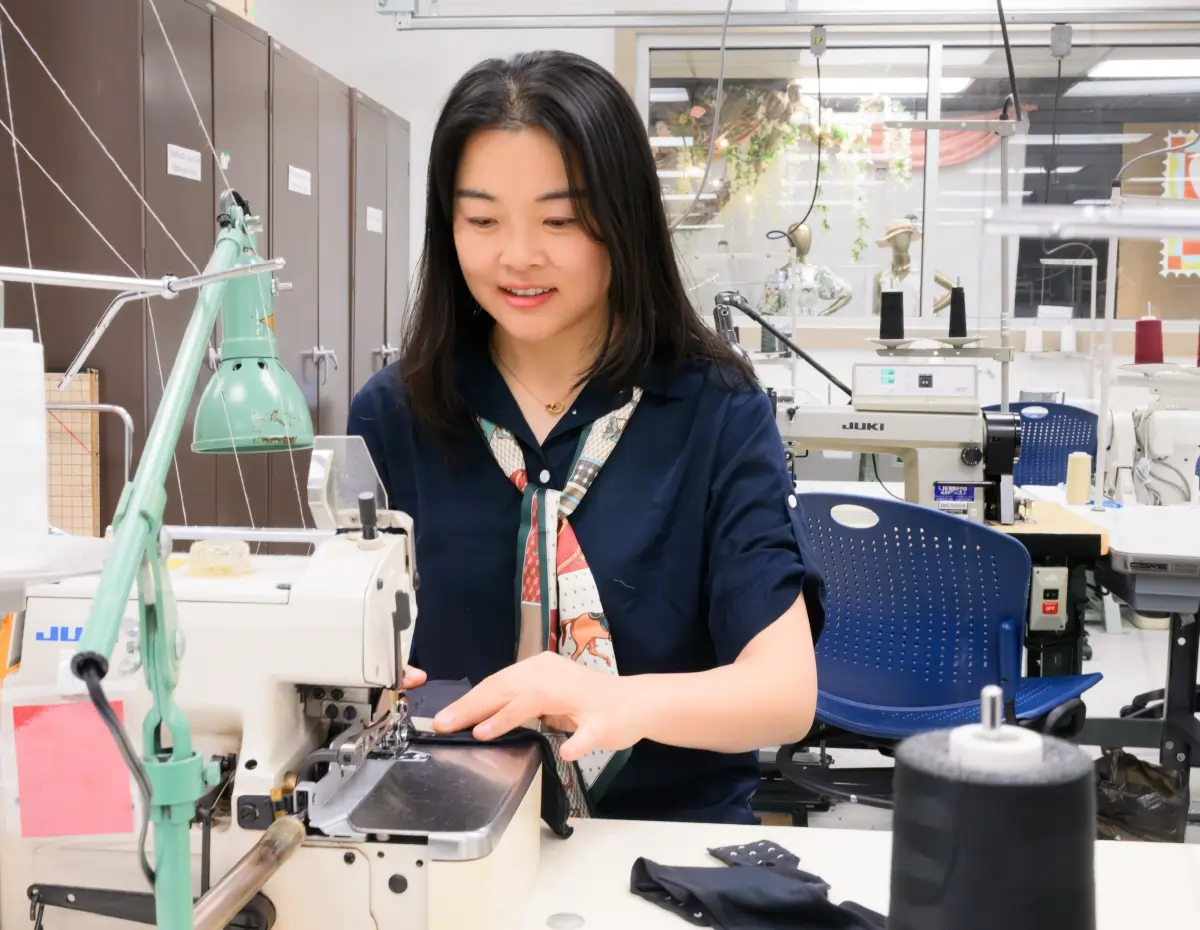 Jia Wu, a woman using an industrial sewing machine; She has on a navy blue shirt and a patterned scarf as she guides black fabric through the machine