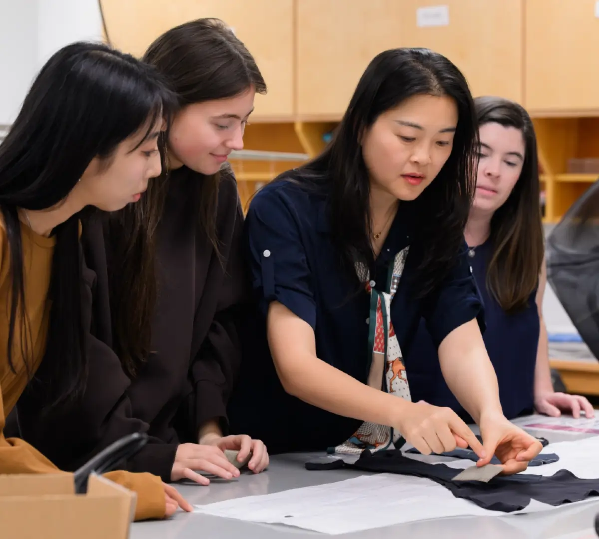 A group of four women are gathered around a work table; Jia Wu is seen pointing to a small fabric swatch being pinned to a dark garment