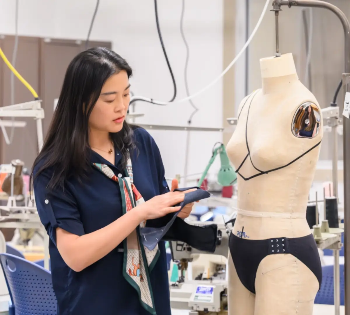Jia Wu, a woman in a navy shirt and patterned scarf holds a piece of dark fabric as she is standing nearby a prototype model dress form