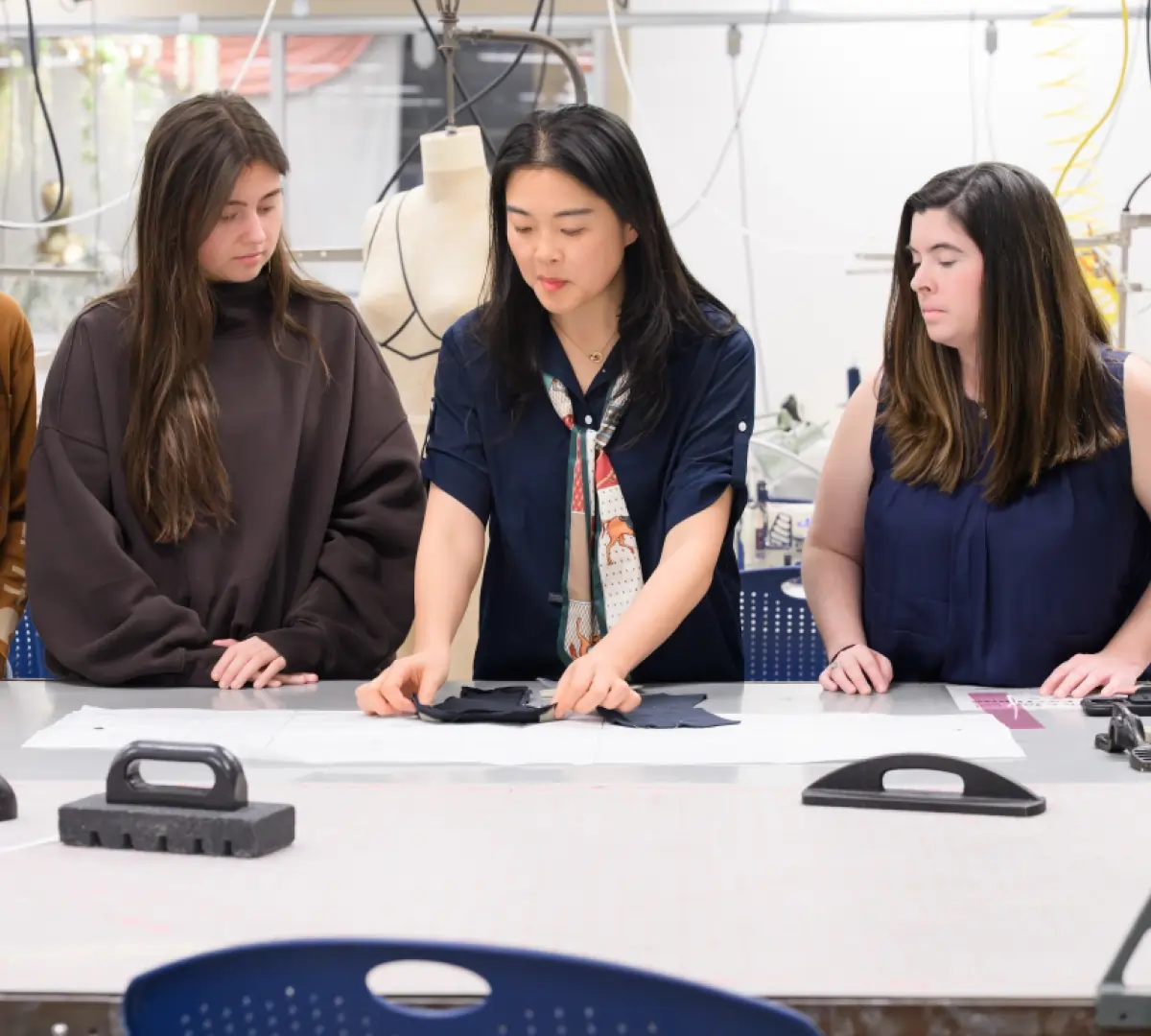 A group of women are gathered around a work table; Jia Wu is seen folding or aligning small fabric swatches being pinned to dark garments